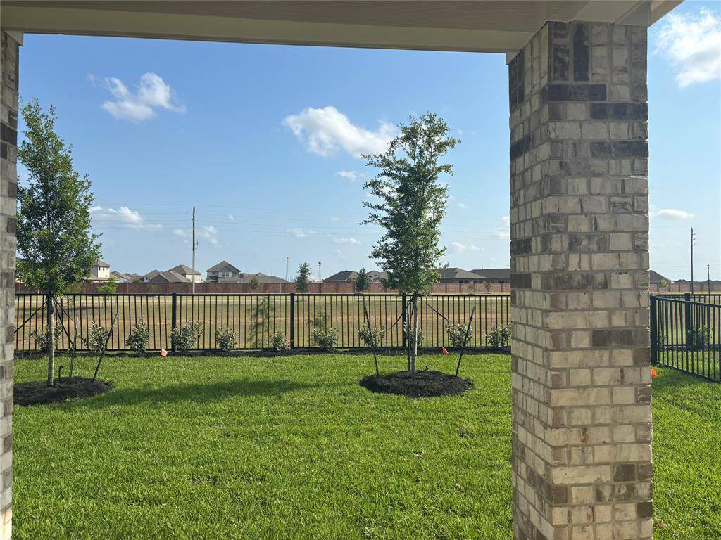 This photo shows a backyard with a covered patio featuring brick columns, a well-maintained lawn, young trees, and a black metal fence. It offers a view of a neighboring area and a clear blue sky.