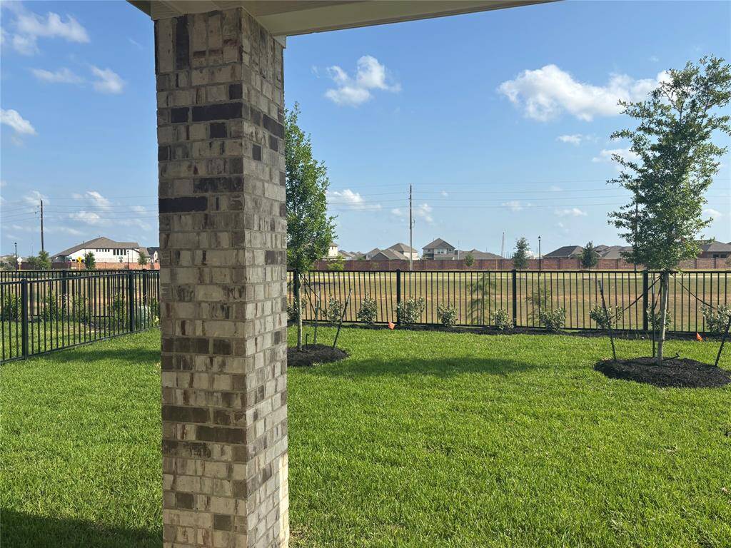 This photo showcases a backyard view from a covered patio, featuring a brick column, well-maintained lawn, young trees, and a black metal fence. The area offers a clear view of neighboring houses and blue skies.