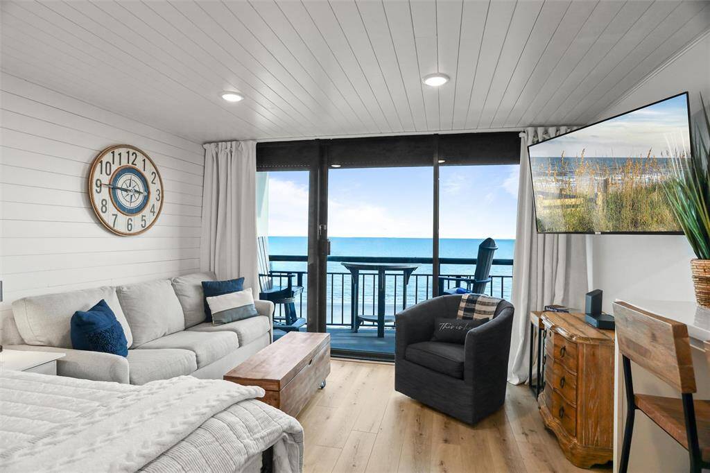 This is a cozy beachfront living room with a clean, nautical theme, featuring a large window that offers an expansive view of the ocean and a balcony for outdoor relaxation.