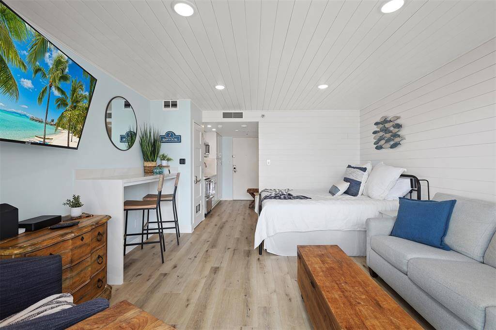 This is a modern, nautically-themed studio with a bright and airy feel, featuring white shiplap walls, light hardwood floors, a space-efficient kitchenette, and a cozy Queen sized sleeping area. The living space is well-appointed with a comfortable queen sleeper sofa allowing space for 4 guests.