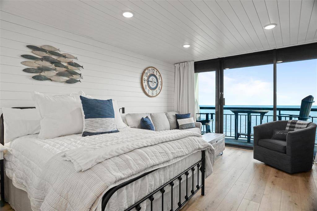 Coastal-themed bedroom with ocean view, featuring a comfortable queen Temperpedic bed, shiplap walls, hardwood floors, and sliding glass doors leading to a balcony.