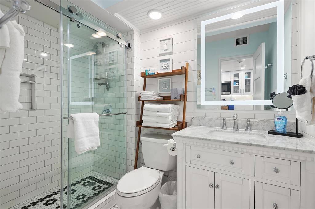This is a modern bathroom featuring a glass-enclosed shower, a large vanity with marble countertop and double sinks, white shiplap walls, wooden shelving for storage, and gorgeus moziac black and white tile. It has a bright and clean aesthetic.
