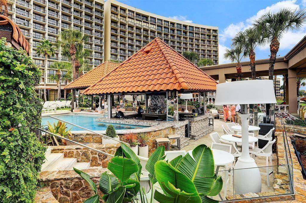 Year round use resort-style pool area is heated in the winter with a poolside bar under a tile-roofed pavilion, surrounded by lush tropical landscaping and tall palm trees, set against the backdrop of the Gulf of Mexico