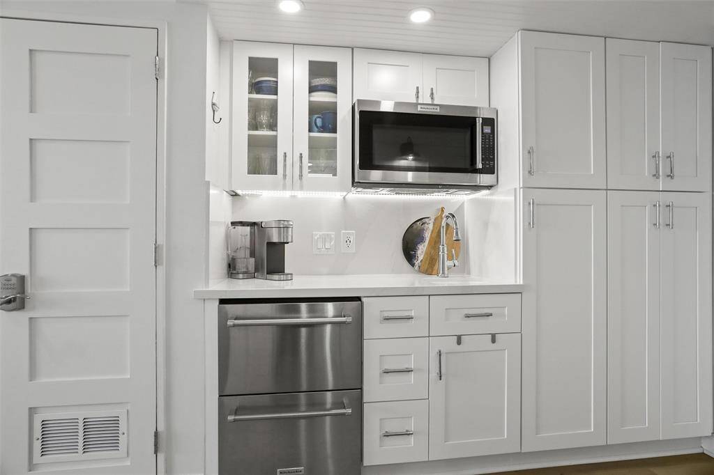 This is a modern, streamlined kitchen with white cabinetry and stainless steel appliances, featuring an under-counter refrigerator, a built-in microwave, and ample storage space. Note that photo is prior to the two burner electric burner being installed. New coffee maker and cooktop not pictured.