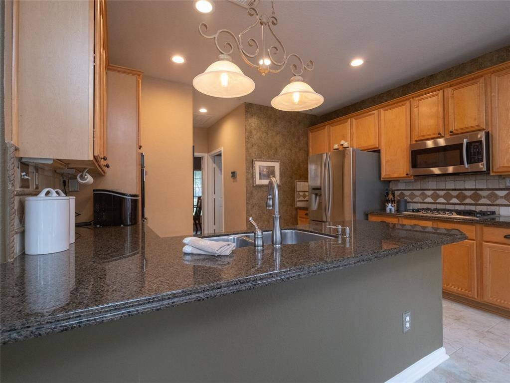 WOW! This kitchen features granite countertops, stainless steel appliances, and wooden cabinetry. The space is well-lit with recessed lighting and a decorative chandelier above the island, creating a warm and inviting atmosphere.