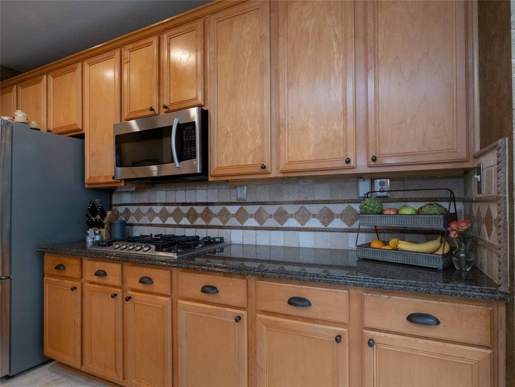 Check out these beautiful warm wood cabinetry, a sleek stainless steel microwave, and a gas cooktop. The granite countertops add elegance, complemented by a decorative tile backsplash.