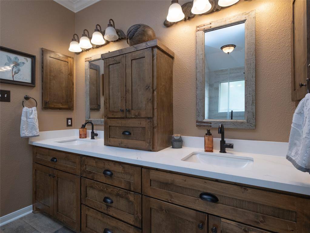 Check out this beautiful and updated primary bathroom! Featuring a dual-sink vanity with rustic wood cabinetry and matching mirrors. It has elegant lighting fixtures and a warm color palette, creating a cozy and functional space.