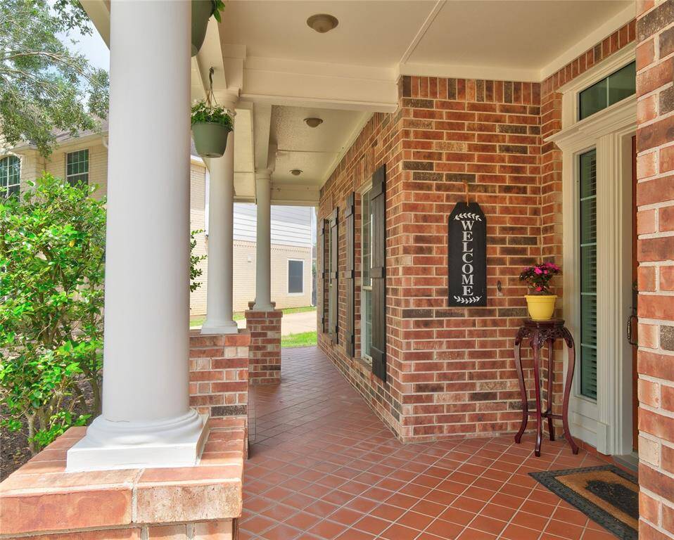 Charming front porch with classic brickwork and white columns, featuring a welcoming sign and potted plant. Ideal for relaxing and enjoying the neighborhood view.
