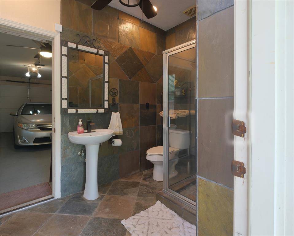 Love this outdoor bathroom with slate tile walls and flooring. It features a pedestal sink, a large framed mirror, and a glass-enclosed shower. The bathroom has an adjoining door leading to a garage, providing convenient access.