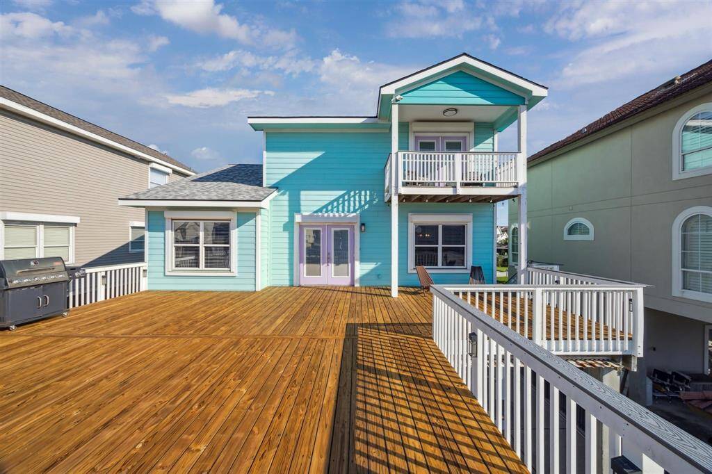 This home features a vibrant turquoise exterior with two levels and a large wooden deck, perfect for outdoor entertaining. It includes a balcony, ample space, and is flanked by neighboring houses, creating a cozy neighborhood feel.