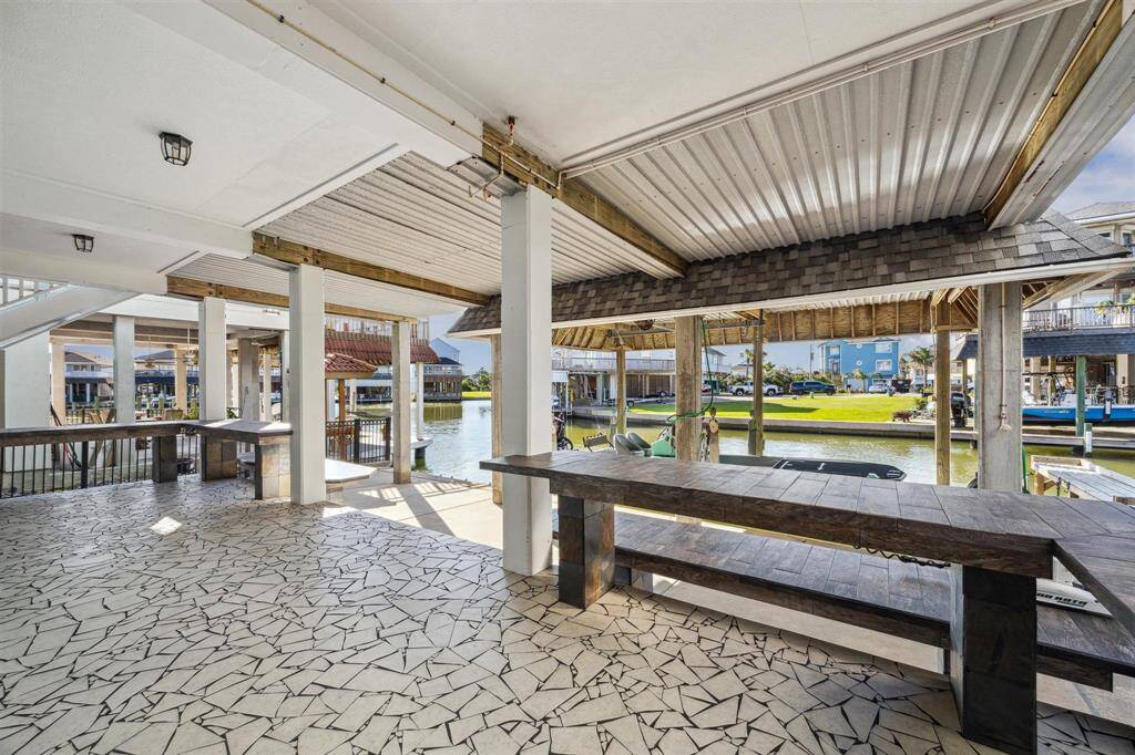 This photo showcases a spacious outdoor patio area with a unique patterned floor, overlooking a serene canal. The space is covered, featuring sturdy wooden beams and ample seating, ideal for relaxation or entertaining with a scenic water view.