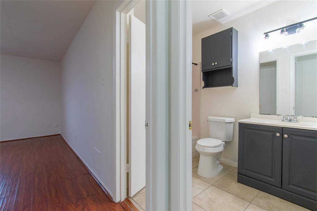 The bathroom features a toilet, a vanity with a dark cabinet, and a large mirror. The adjacent room has wood-style flooring, offering a versatile space.