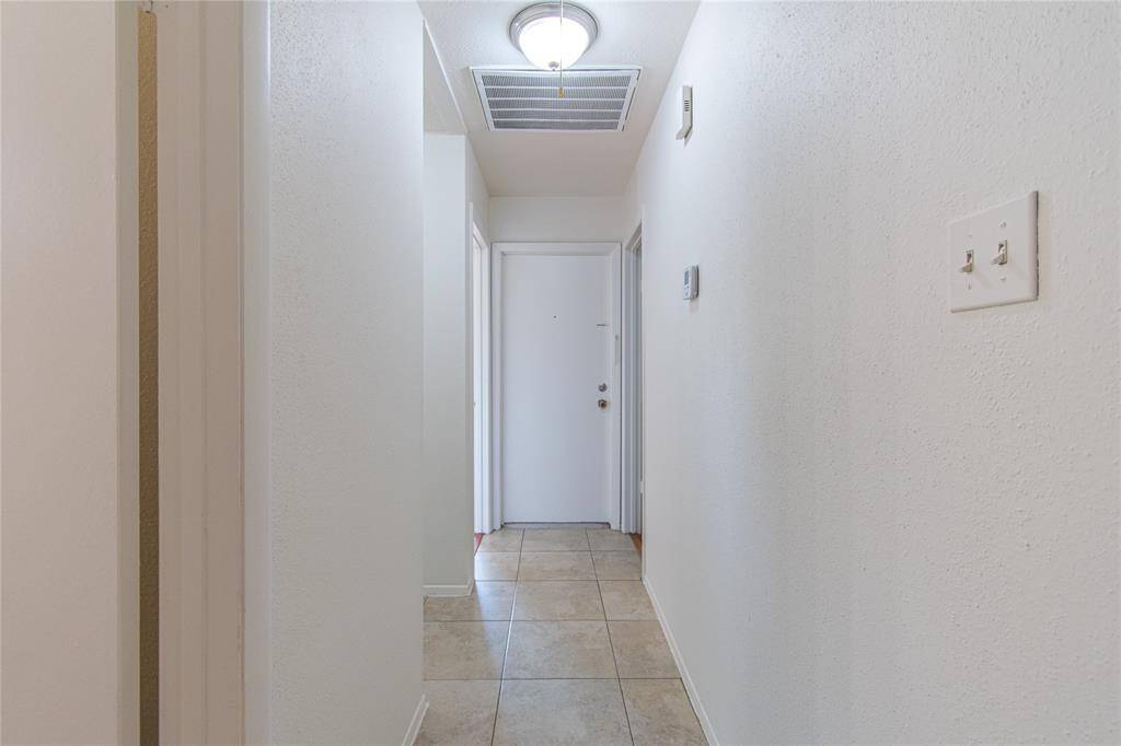 This photo shows a clean, narrow hallway with tiled flooring, white walls, and a ceiling light fixture. At the end of the hallway is a door, suggesting access to another room or entrance.