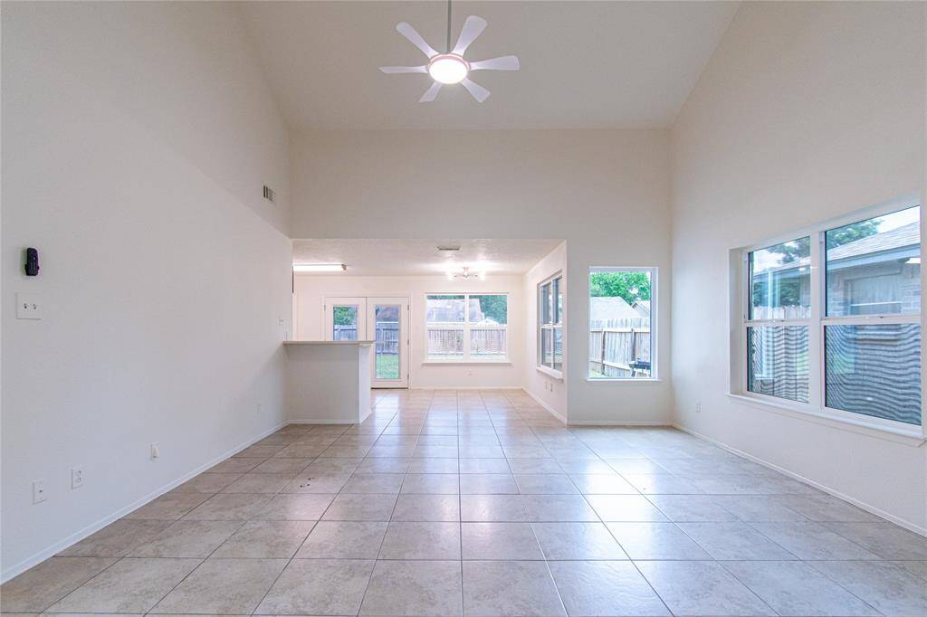 This bright, open-concept living space features high ceilings, tile flooring, and large windows that bring in ample natural light. A ceiling fan and neutral walls add a modern touch, while the layout seamlessly connects to a kitchen area with views of the fenced backyard.