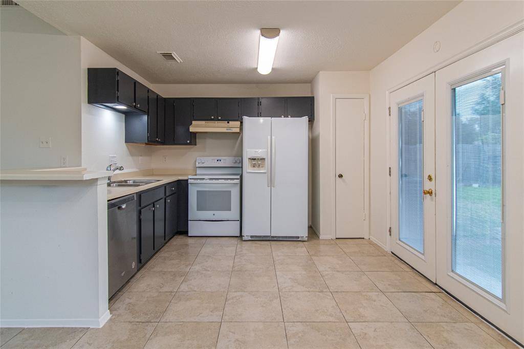 This kitchen features dark cabinets, a white fridge, and stove, with ample counter space. It has tiled flooring and double glass doors leading to the outside, providing natural light and easy access to the backyard.