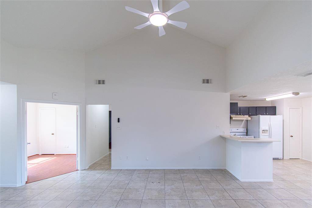 Bright, open living space with high ceilings and tile flooring. The room features a ceiling fan and connects to a kitchen with a breakfast bar and modern appliances. Adjacent rooms offer additional space with natural light.