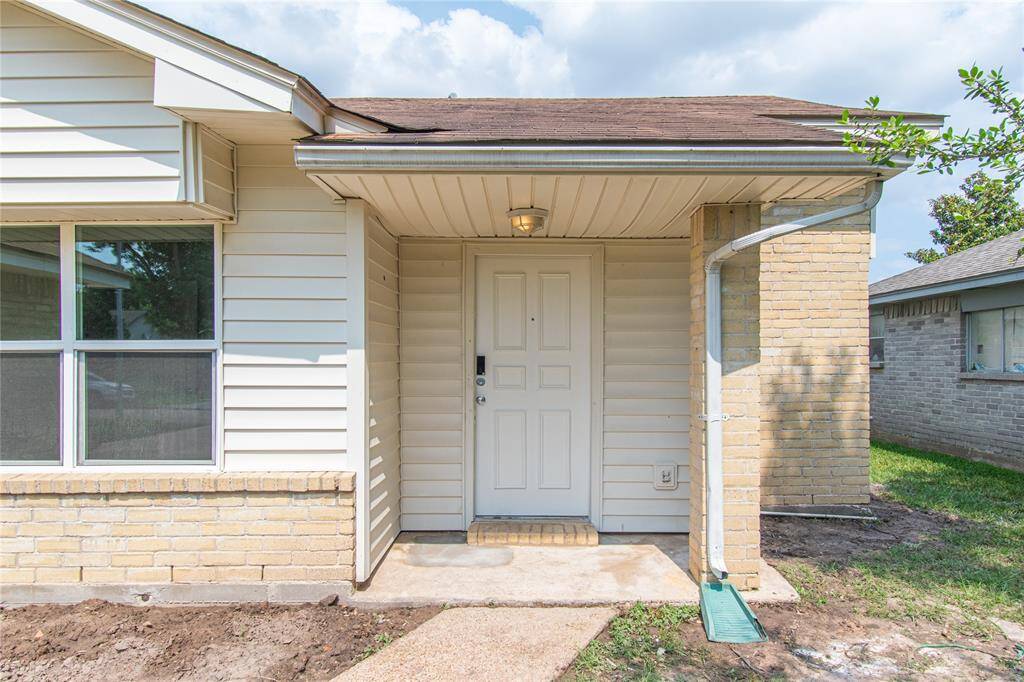 Front entrance of a home, featuring a covered porch area with a white door, and brick accents. The pathway leads to a small, well-maintained yard.