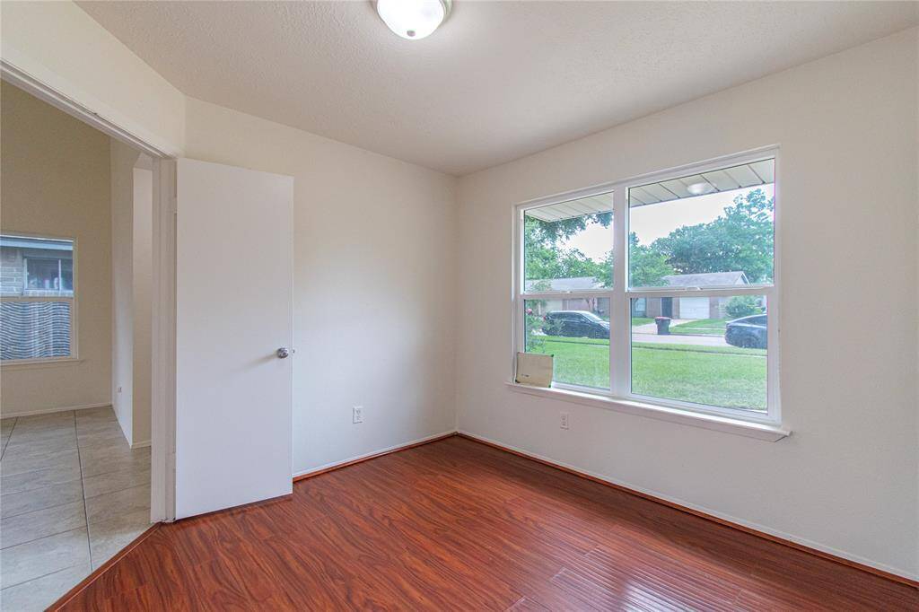 This room features laminate wood flooring, neutral walls, and a large window offering a view of the front yard and ample natural light. It connects to a tiled hallway.