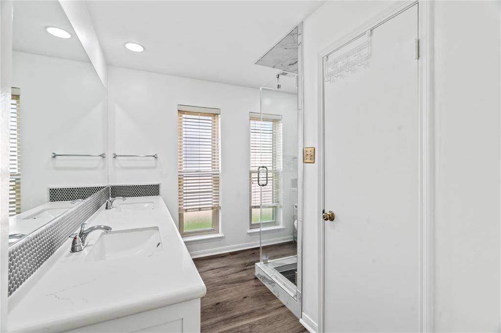 Bright, modern bathroom featuring a dual-sink vanity, large mirror, glass-enclosed shower, and wood-style flooring. Two windows provide natural light, enhancing the clean white decor.