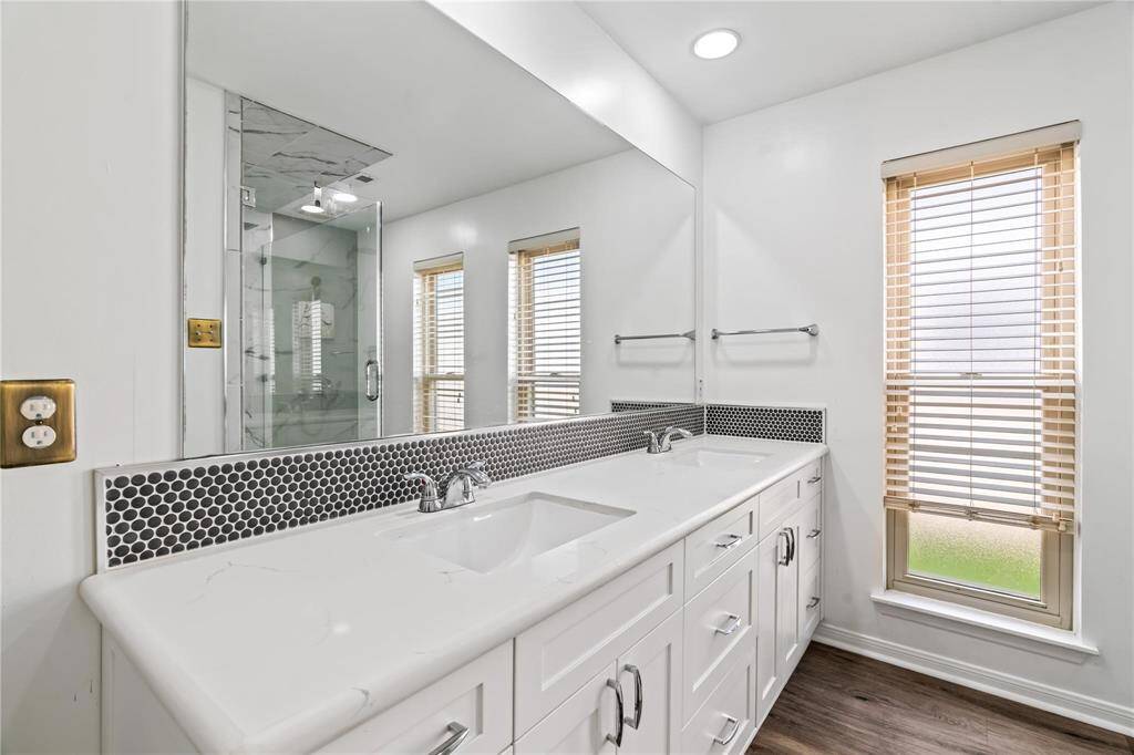 Modern bathroom featuring a double vanity with marble countertops, sleek faucets, and a stylish tile backsplash. The space is bright with large windows, wooden blinds, and a glass-enclosed shower.