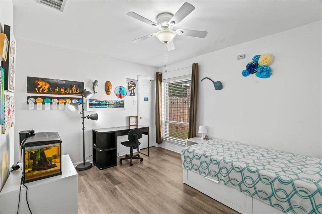 This bright bedroom features a modern design with wood flooring, a ceiling fan, and ample natural light from a large window. It includes a single bed, a desk with a chair, and various wall decorations, creating a cozy and functional space.