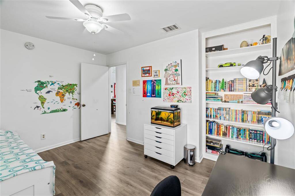 This room features a modern, cozy design with laminate wood flooring and white walls. It includes built-in shelves filled with books and decor, a desk with a lamp, and a small aquarium. The wall is adorned with colorful artwork and a world map, providing a vibrant and inviting atmosphere.