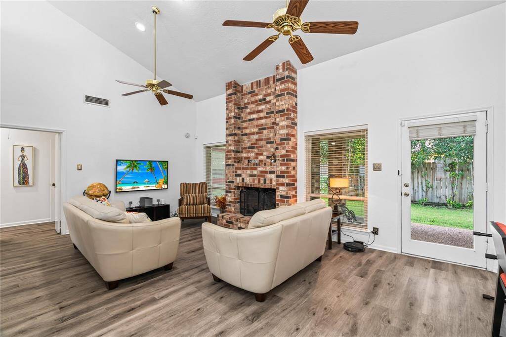 This inviting living room features high ceilings with two ceiling fans, a central brick fireplace, and ample natural light from large windows and a glass door leading to the backyard. The space has a warm, open feel with light wood flooring and comfortable seating.