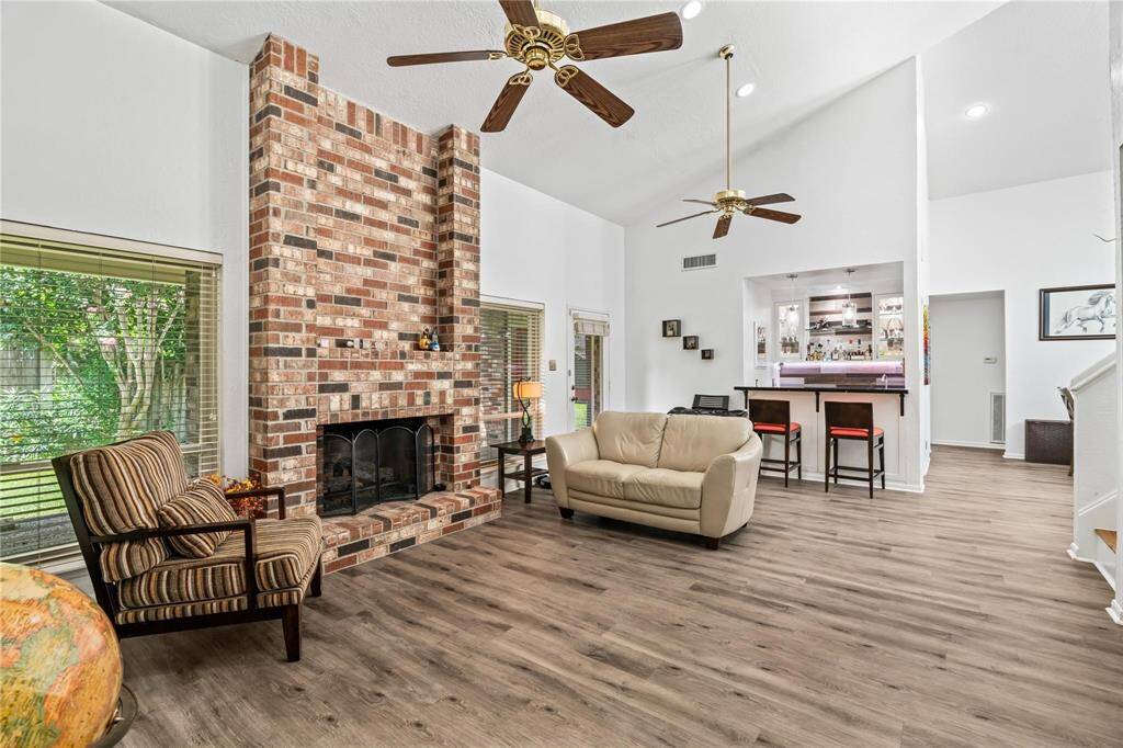 This cozy living room features a high ceiling with ceiling fans, a charming brick fireplace, and large windows for natural light. The open layout includes a bar area, perfect for entertaining.