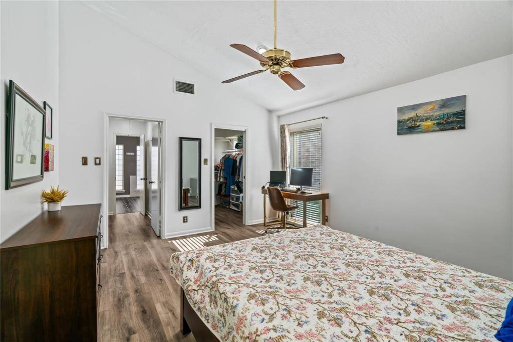 This bright bedroom features a vaulted ceiling with a ceiling fan, wood flooring, and large windows for natural light. It includes a floral-patterned bedspread, a desk area, and access to a walk-in closet and en-suite bathroom.