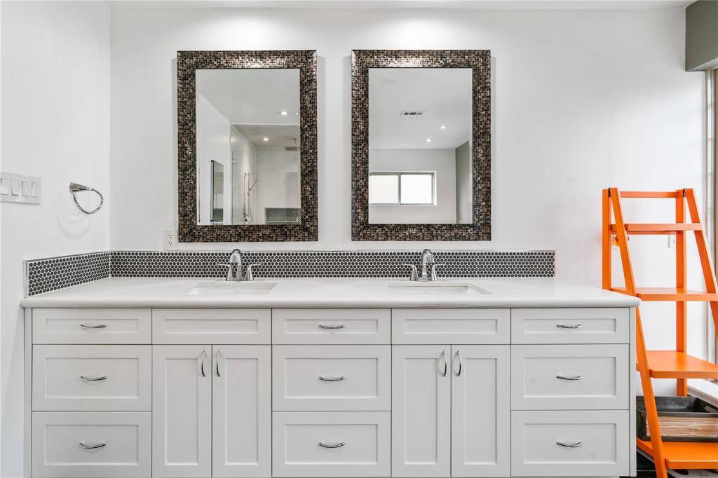 This bathroom features a modern double vanity with white cabinetry, dual sinks, and stylish mosaic-framed mirrors. The space is accented by a unique orange shelving unit, offering a pop of color and additional storage.