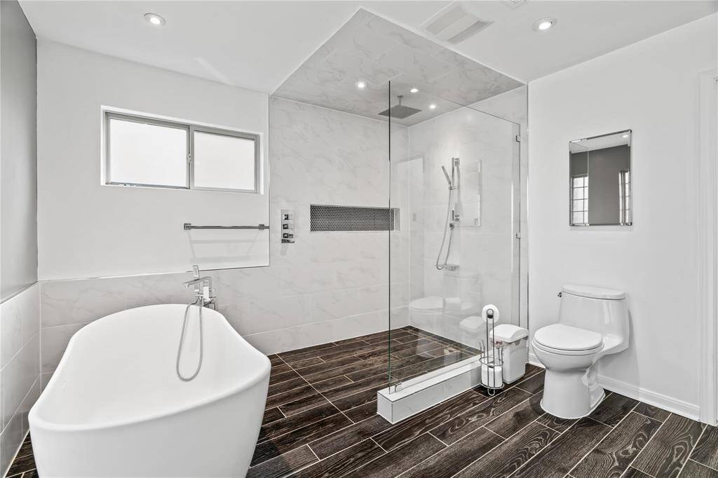 This modern bathroom features a sleek, freestanding bathtub, a glass-enclosed shower, and elegant dark wood-style flooring. Bright and spacious, it offers a clean, minimalist design with ample natural light.