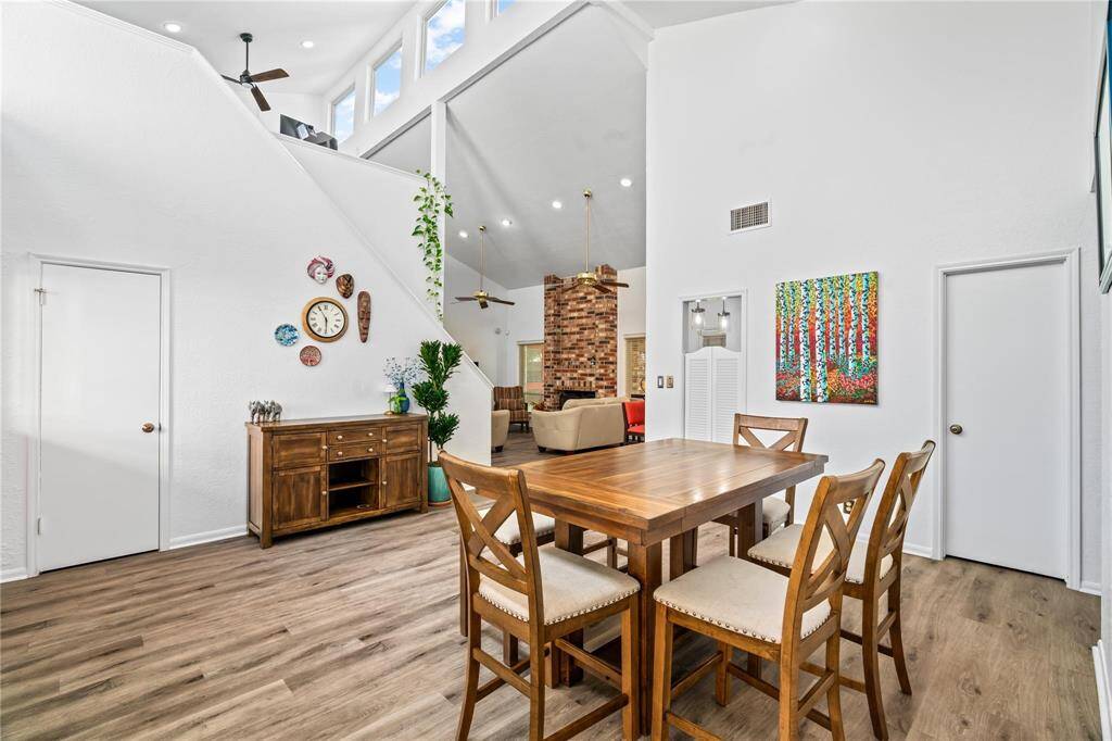 This spacious, bright dining area features high ceilings, wood flooring, and modern lighting. It opens to a cozy living space with a brick accent wall, creating a warm and inviting atmosphere.