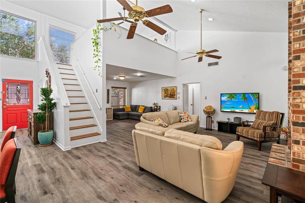 Spacious living area with high ceilings, featuring a cozy sectional and a bright, inviting atmosphere. The open staircase leads to a loft, and large windows provide plenty of natural light. A brick fireplace adds warmth, and the red front door offers a pop of color. Ideal for comfortable family living.