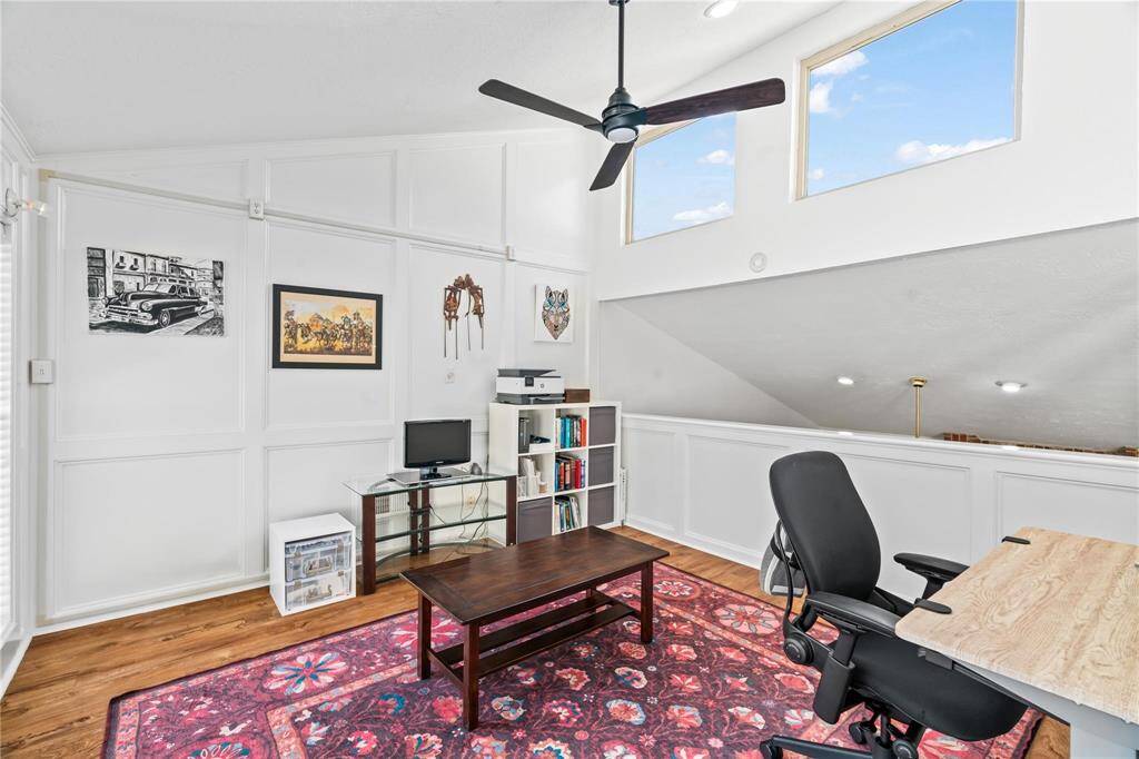 This bright loft space features high ceilings with skylights, a ceiling fan, and wood flooring. It includes a workspace with a desk and chair, a cozy setup with a rug and coffee table, and built-in shelving. Ideal for a home office or creative studio.