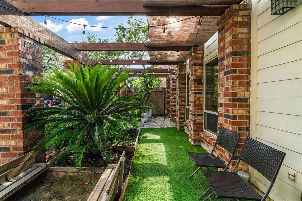 Charming outdoor space featuring a pergola with string lights, lush greenery, and cozy seating. The area is bordered by attractive brickwork and siding, creating a welcoming and private ambiance perfect for relaxation.