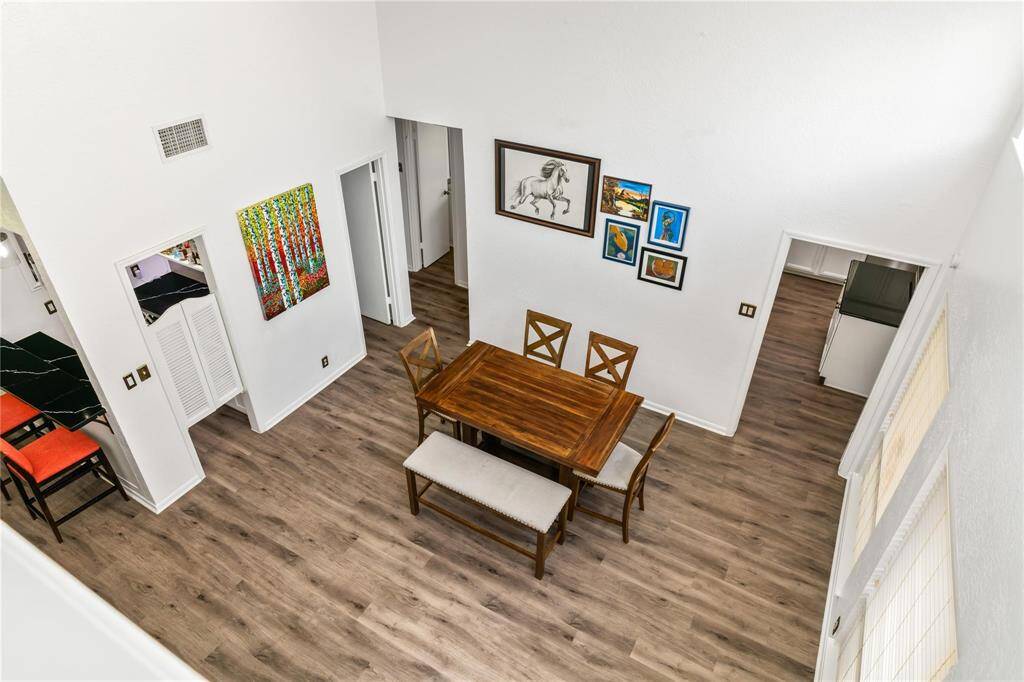 This photo showcases a bright, open dining area with high ceilings and wood flooring. The room features a wooden dining set and is adorned with colorful artwork. Adjacent spaces, including a kitchen and hallway, are visible, enhancing the home's spacious feel.
