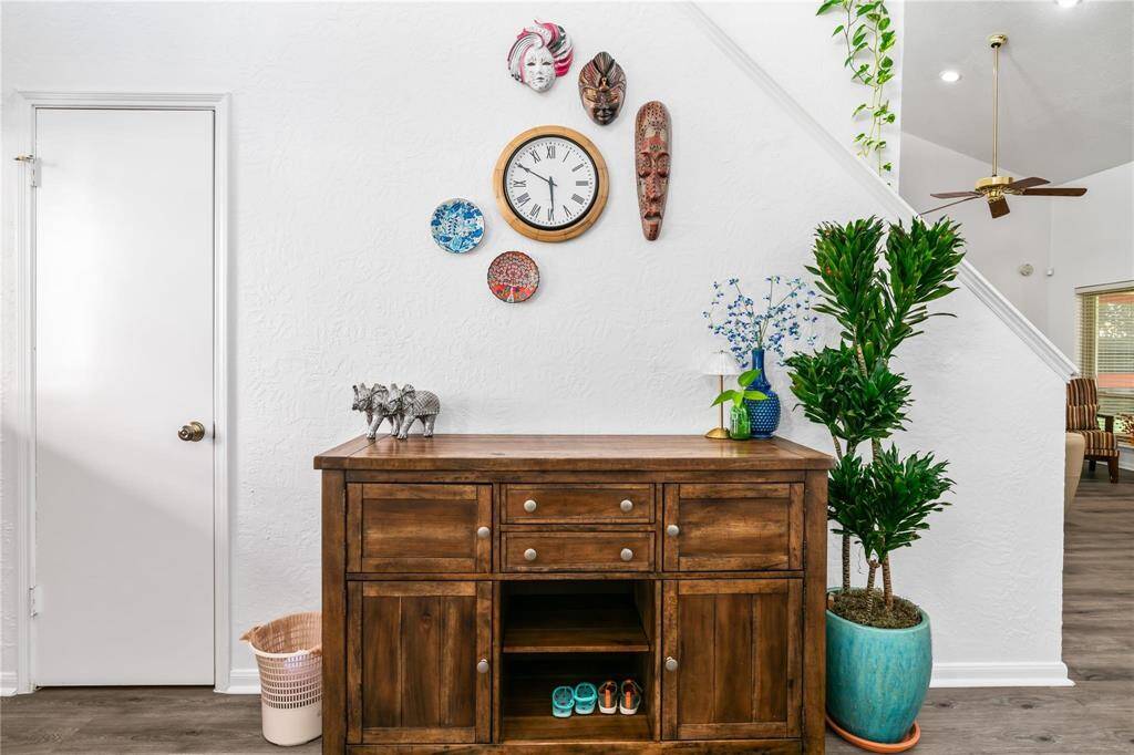 This inviting entryway features a textured white wall adorned with eclectic decor and a clock. A wooden console table offers storage and display space, complemented by a potted plant and decorative items. The area has a cozy, welcoming feel with a view into the living space.
