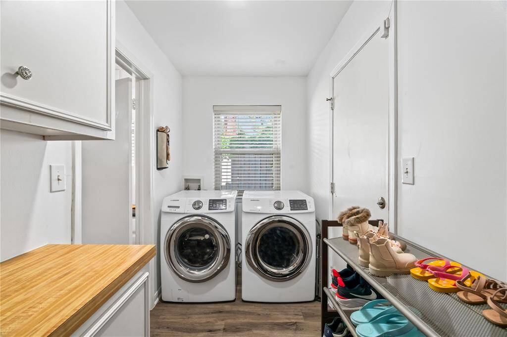 This laundry room features modern front-loading washer and dryer units, a window for natural light, white cabinetry, and a wooden countertop. It offers ample space for organization, with a shoe rack and practical layout.