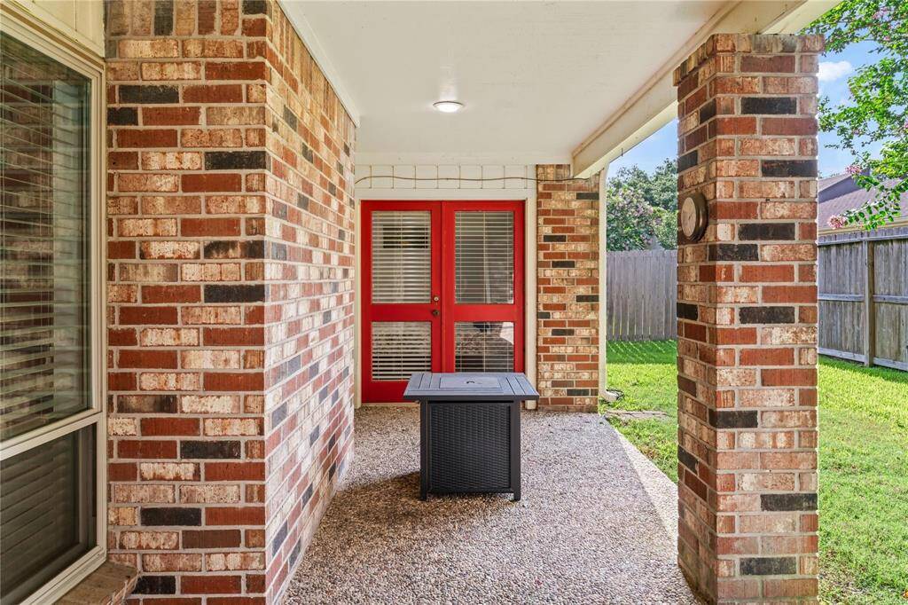 This photo showcases a cozy covered patio with brick pillars and red French doors, opening to a fenced green backyard, perfect for outdoor relaxation and entertaining.