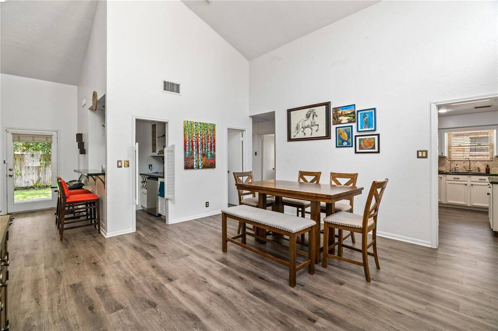 This photo showcases a bright dining area with high ceilings and wood flooring. The space features a wooden dining set with seating for six and a bar area with red stools. Artwork adorns the walls, and the room connects to a kitchen with white cabinetry. A door leads to a fenced backyard, adding natural light.
