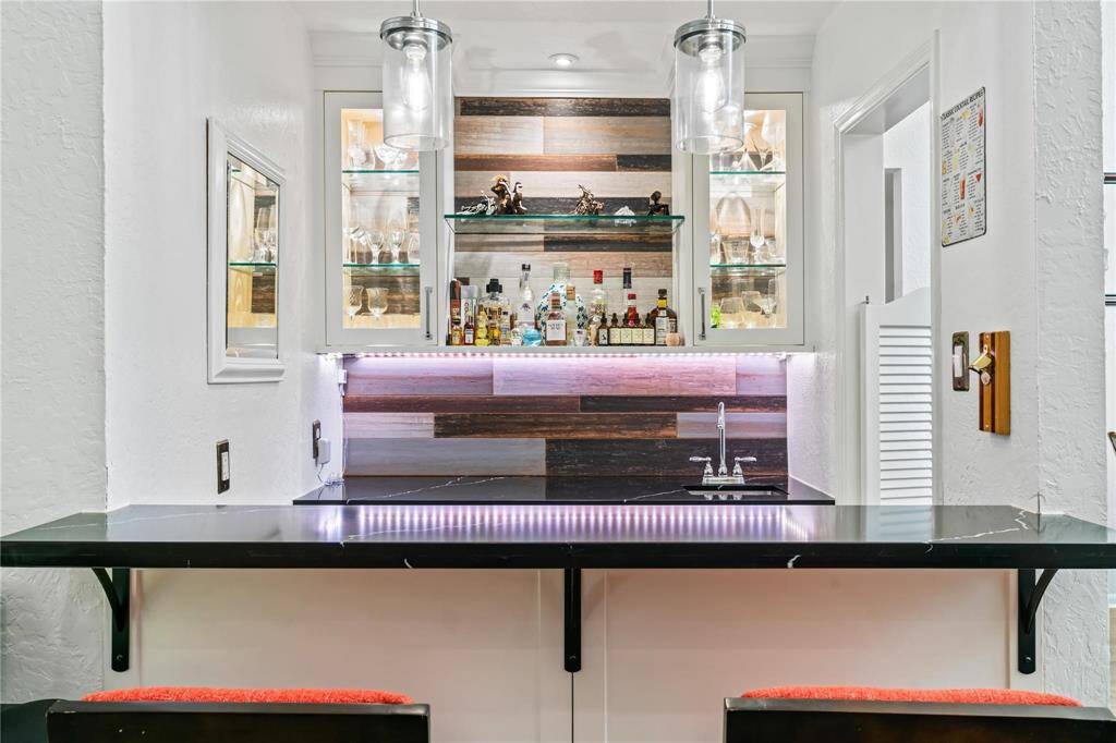 This home features a stylish wet bar with a sleek black countertop, modern pendant lighting, and glass cabinets for displaying glassware. The backsplash has a contemporary wood panel design, and there's ample space for entertaining.