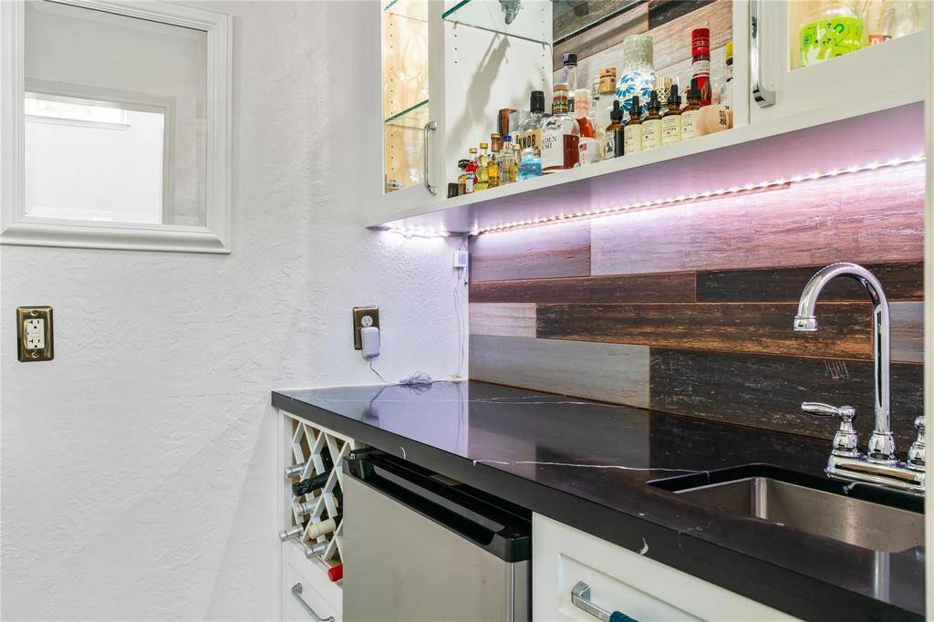 This photo showcases a modern home bar area with a sleek black countertop, stainless steel sink, and a stylish backsplash of mixed wood tones. It features under-cabinet lighting, a small wine rack, and a mini fridge, creating a perfect space for entertaining.