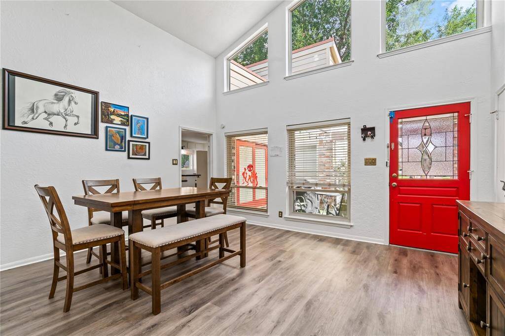 This bright dining area features high ceilings and large windows, allowing ample natural light. The space is enhanced by a vibrant red front door, modern wood flooring, and tasteful wall art, creating a welcoming and stylish atmosphere.