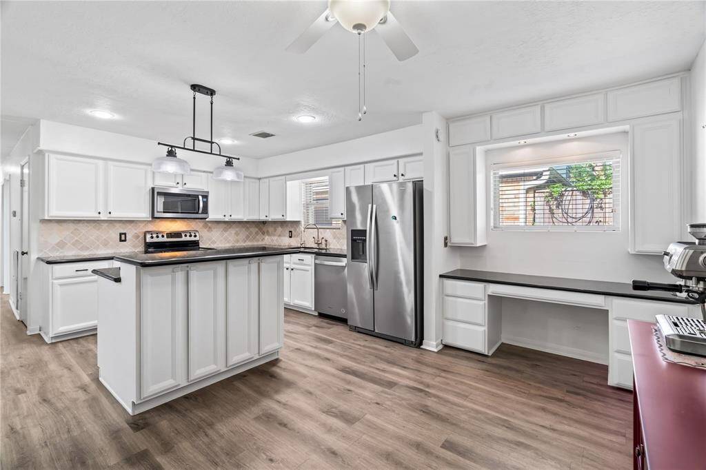 This modern kitchen features sleek white cabinetry, stainless steel appliances, and a center island with pendant lighting. The spacious layout includes ample counter space and a built-in desk area, complemented by wood-style flooring and a large window for natural light.