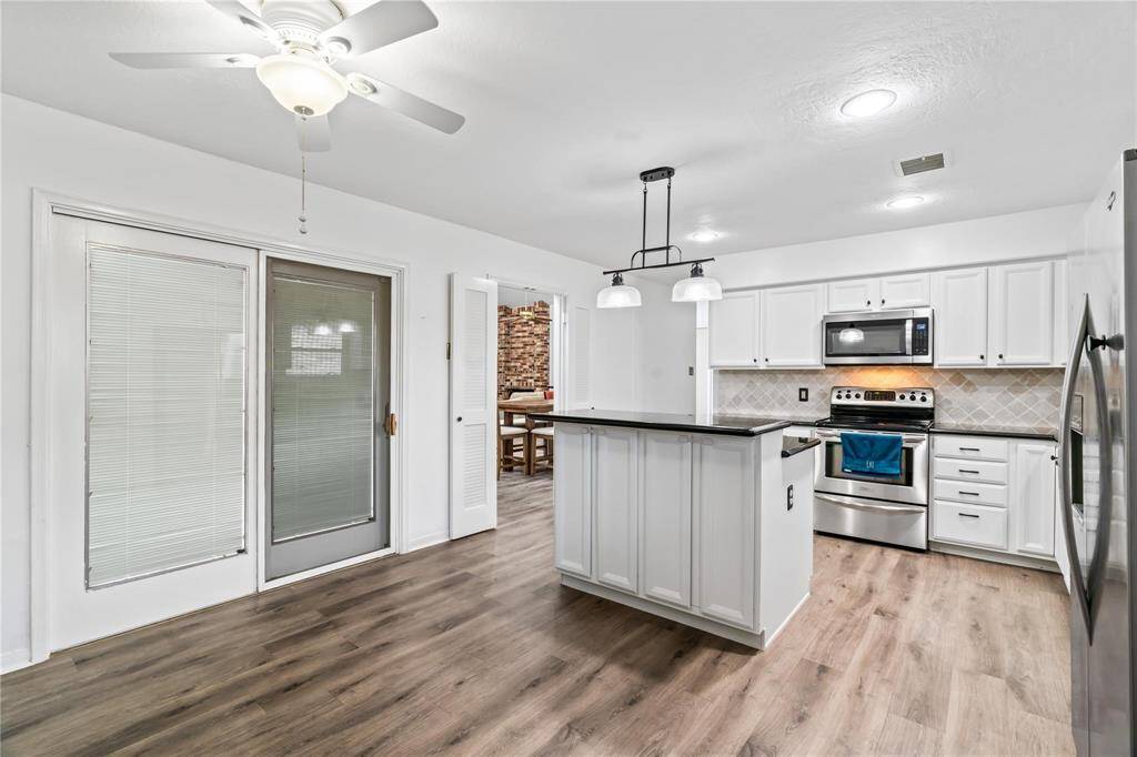 This photo showcases a modern kitchen with white cabinetry, stainless steel appliances, and a central island. The space features wood-look flooring, a ceiling fan, recessed lighting, and a sliding glass door leading outside.