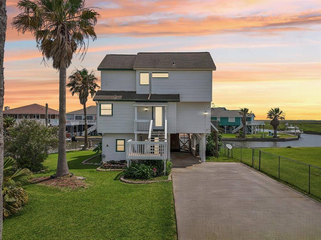 This is a charming elevated white house with a welcoming front balcony, surrounded by lush green grass and a few palm trees. The property features a spacious driveway and is situated in a serene, open area with a clear blue sky backdrop.