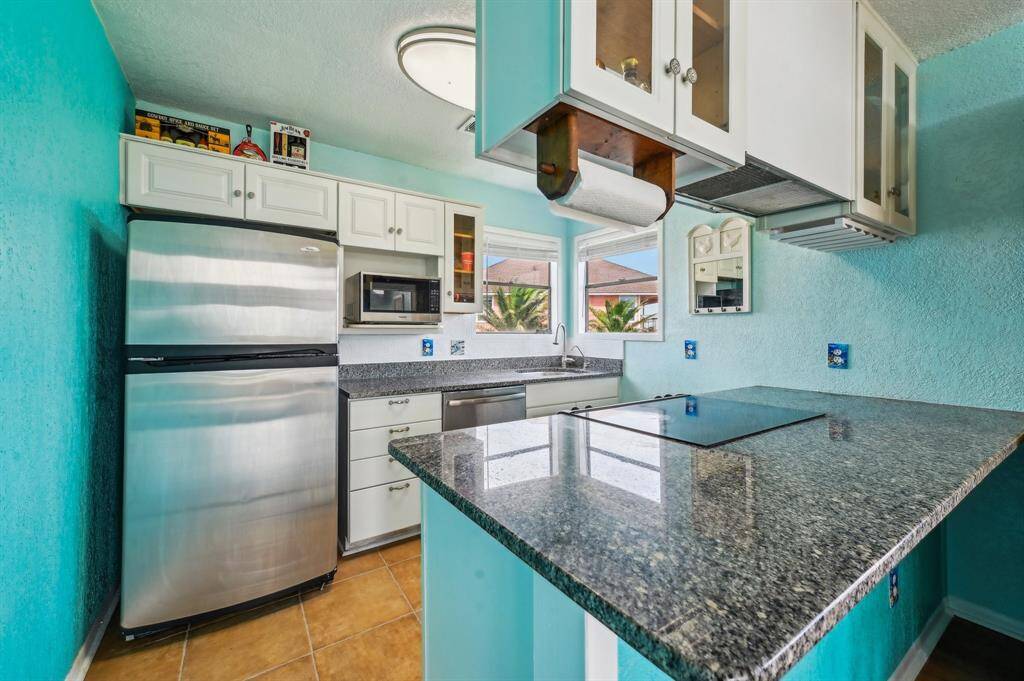 This bright, inviting kitchen features turquoise walls, hardwood floors, and ample natural light from large windows. It includes modern stainless steel appliances, a stylish chandelier, and a cozy dining area with padded stools.