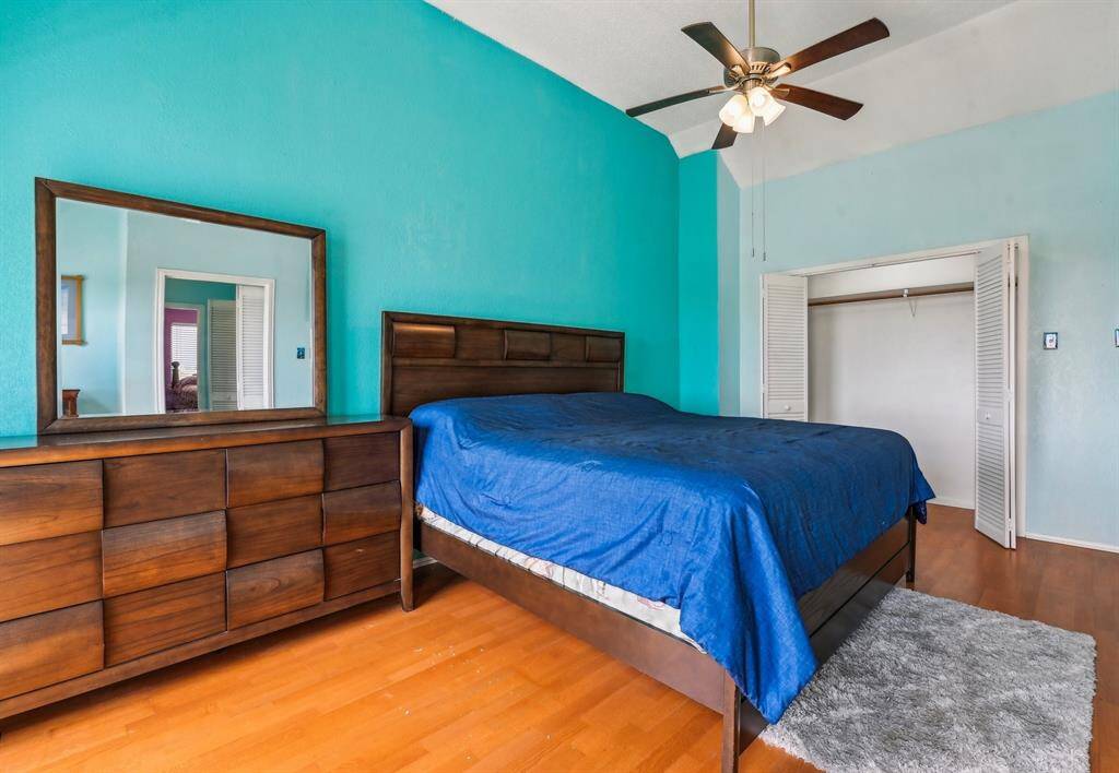 This photo shows a hallway with light blue walls and wood flooring, leading to a bedroom. The hallway features white louvered closet doors on the right.