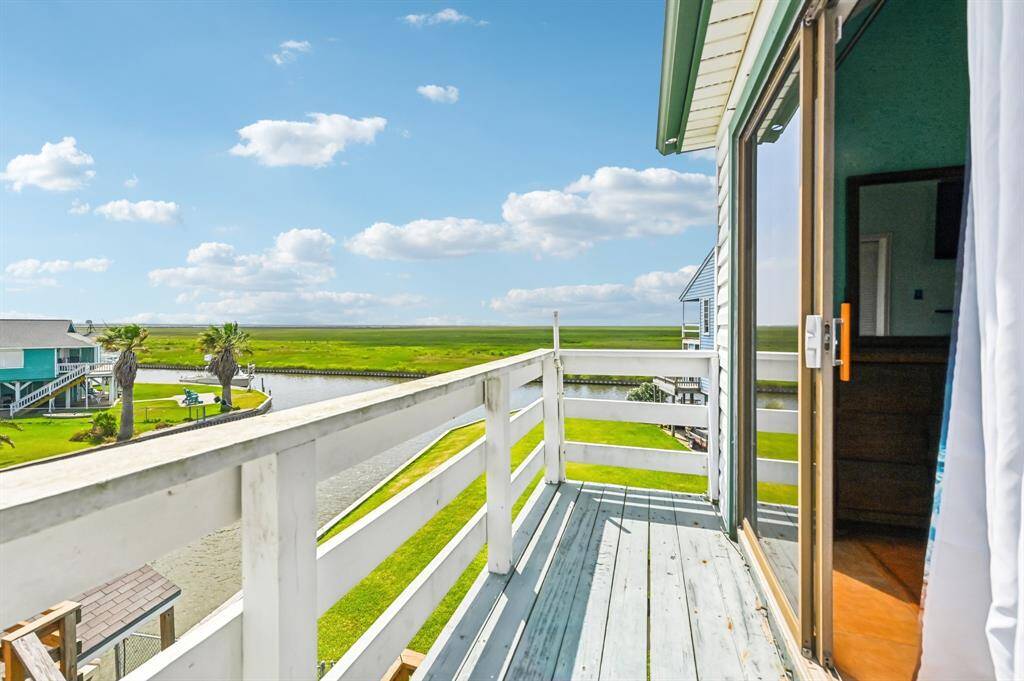 This photo showcases a spacious outdoor deck with artificial turf, perfect for relaxation or entertaining. It features a wooden railing, a small table with chairs, and open views of greenery and a distant house, under a bright blue sky with scattered clouds.