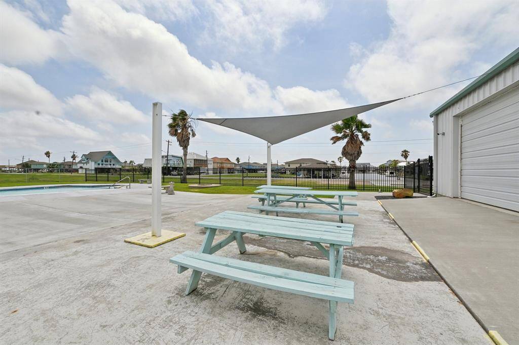 This photo features a well-maintained outdoor pool area with a clear blue swimming pool, surrounded by concrete decking. Adjacent to the pool is a small light-colored building with seating and storage space. The area is enclosed by a fence, with palm trees and residential homes visible in the background, creating a relaxed and inviting atmosphere.